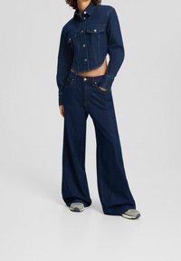 Mörkblå denimjacka i kort modell med knäppning och två framfickor, tillsammans med löst sittande mörkblå jeans. Subtila sömnadsdetaljer.