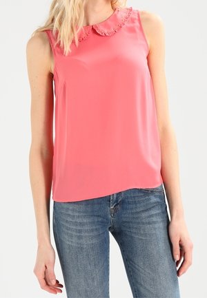 Mujer con blusa sin mangas de color coral rosado con cuello con volantes y jeans de mezclilla azul, vista desde los hombros hasta las caderas.