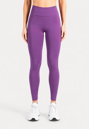 Femme portant des leggings violets taille haute et des baskets blanches, debout droite devant un fond blanc uni.