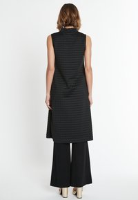 Schwarzes ärmelloses Kleid mit einem strukturierten, gesteppte Muster, das einen hohen Kragen und einen lockeren Schnitt aufweist, kombiniert mit weit geschnittenen schwarzen Hosen und goldenen Schuhen.