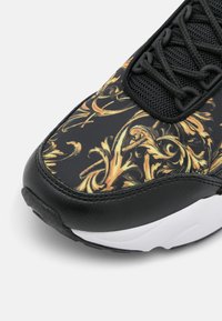 Svart sneaker-tå med intrikata blomster swirl-mönster i guld och orange, svarta snörningar, mesh-tunga och vita detaljer på sulan.