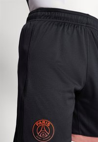 Mustat urheilushortsit, jotka on valmistettu teksturoidusta kankaasta. Alhaalla on oranssi Paris Saint-Germain -logo. Mukava joustava vyötärö.