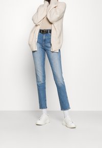 Krämfärgad kavaj över en beige topp, högmidjade ljusblå jeans och vita sneakers. Har ett strukturerat mönster och ett svart bälte.