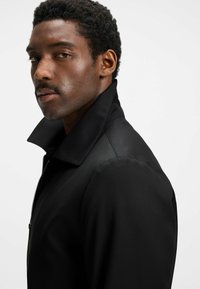 Schwarze Jacke mit hohem Kragen und Reißverschluss vorne. Glattes Material mit sichtbaren Nähten, taillierte Form und ohne sichtbare Muster oder Hardware.