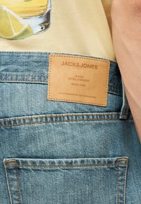 Κοντινή λήψη της πίσω τσέπης μπλε τζίν με ένα ταμπελάκι από μαύρο δέρμα με την επιγραφή "JACK & JONES" και ένα κίτρινο πουκάμισο με λεμονοτυπία.