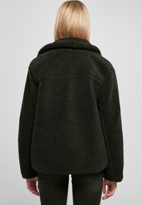 Mørkegrøn, blød sherpa-stil jakke med krave og en afslappet pasform. Bagfra ses glatte syninger og en let buet kant.