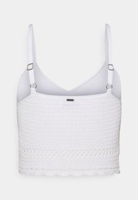 Witte cropped top met geweven textuur, verstelbare bandjes en gegolfde onderrand. Heeft een v-halslijn en een klein metallic logo detail.