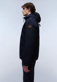 Junger Mann im Profil, der eine marineblaue Kapuzenjacke mit einem runden Logo-Patch auf dem Oberarm trägt, vor einem hellgrauen Hintergrund.