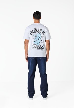 Άνδρας στέκεται με την πλάτη γυρισμένη φορώντας λευκή μπλούζα με μπλε ζάρια και το κείμενο "California Feeling Lucky", συνδυασμένη με σκούρο μπλε τζιν και λευκά παπούτσια.