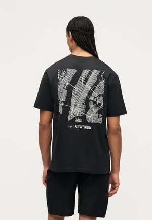 Person med flettet hår vendt væk, iført en sort T-shirt med detaljeret hvidt gade kort over New York på ryggen.