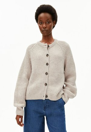 Personne aux cheveux courts et bouclés portant un cardigan beige clair côtelé boutonné et un jean bleu, debout avec une main dans la poche.