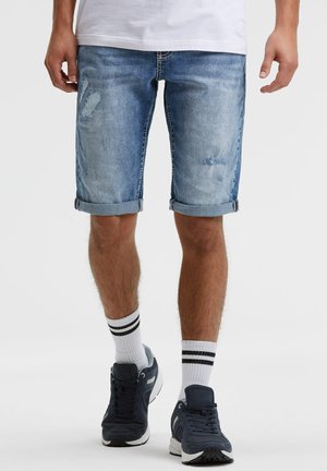 Mann trägt hochgekrempelte, abgenutzte Jeansshorts, weiße Crew-Socken mit schwarzen Streifen und dunkelblaue Sneaker und steht vor einem schlichten Hintergrund.