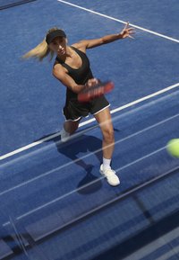 Tenisový hráč v černém sportovním oblečení s kšiltem, máchající raketou na modrém kurtu s bílými čarami, soustředěný na žlutý tenisový míček.