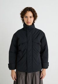 Schwarze isolierte Jacke mit hohem Kragen, Fronttaschen und weiten Ärmeln. Aus einem strukturierten Stoff für Wärme und Komfort gefertigt.