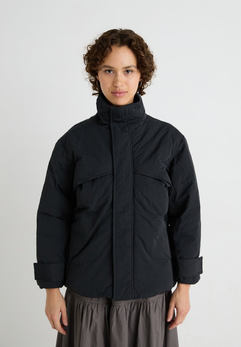 Schwarze isolierte Jacke mit hohem Kragen, Fronttaschen und weiten Ärmeln. Aus einem strukturierten Stoff für Wärme und Komfort gefertigt.