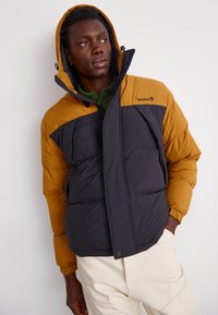 Pufjacke i sort og sennepsgul med hætte, lynlås og elastiske manchetter; quiltet tekstur med et logo på brystet.