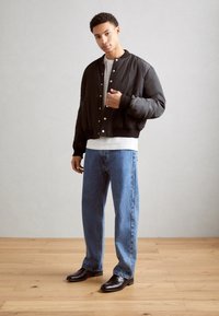 Veste bomber noire avec des boutons-pression, portée sur un t-shirt blanc, associée à un jean bleu clair et des chaussures noires brillantes, se tenant à l'intérieur.