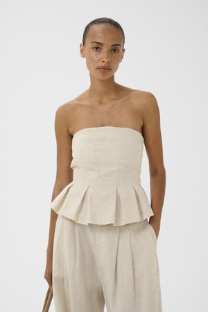 Femme aux cheveux tirés en arrière portant un top beige plissé sans bretelles et un pantalon large assorti sur fond uni, tenant un sac.