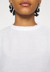 Haut blanc texturé avec un col à volants, fabriqué dans un tissu léger et respirant. Design simple, sans motifs ni accents supplémentaires.