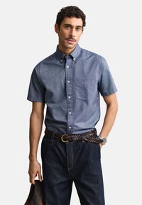 Mann trägt ein kurzärmeliges blaues Hemd mit Knöpfen, dunkle Jeans mit geflochtenem braunem Gürtel, Armbanduhr, hält einen dunkelbraunen Gegenstand.
