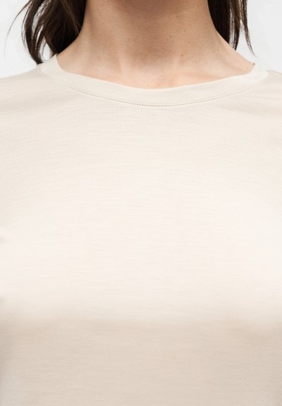 Κοντινό πλάνο ενός ατόμου που φοράει μια απλή μπεζ μπλούζα με στρογγυλή λαιμόκοψη, δείχνοντας τον λαιμό και το πάνω μέρος του στήθους.