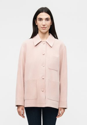 Mujer con cabello largo y oscuro que lleva una chaqueta rosa claro con botones y tres bolsillos delanteros, de pie frente a un fondo blanco liso.