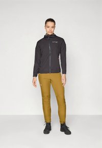 Črno lahko jakno s kapuco, z zadrgo spredaj in rdečimi poudarki, kombinirano s slim krojenimi rumenimi hlačami in črno obutvijo.