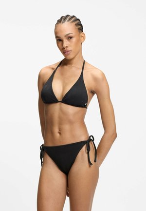 Kobieta z warkoczami na włosach, ubrana w czarne teksturowane bikini z topem typu halter i majtkami z wiązaniem po bokach, stojąca na białym tle.