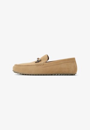 Tan suede moccasin with a rounded toe, decorative stitching, and a small silver accent. Flat rubber sole with textured grip.