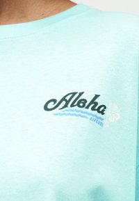 Φωτεινό γαλάζιο πουκάμισο με το κείμενο "Aloha", σχέδιο μπλε κύματος, μικρό γραφικό με λουλούδια και το όνομα της μάρκας "Rip Curl" κοντά στην κλείδα.