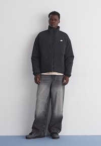 Svart, zip-up pufferjacka med hög krage, bärs över en ljus skjorta. Kombinerad med bleka gråa baggy jeans och mörka skor. Enkla design detaljer.
