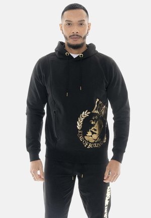 Mann trägt schwarzen Hoodie und Hose mit goldenem "King of Boxing" Löwenlogo, steht vor hellem einfarbigem Hintergrund.