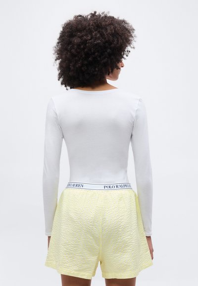 Άτομο με σγουρά μαλλιά φοράει λευκό μακρυμάνικο μπλουζάκι και κίτρινα υφασμάτινα σορτς με ζώνη Polo Ralph Lauren, με την πλάτη προς τα εμάς.
