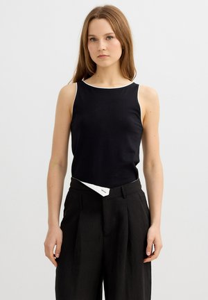 Mujer joven con cabello largo castaño claro, vistiendo una camiseta negra sin mangas y pantalones negros de tiro alto y piernas anchas sobre un fondo liso.