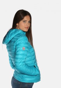 Doudoune turquoise avec capuche, présentant un design matelassé, des poches latérales et un patch rond sur la manche gauche.