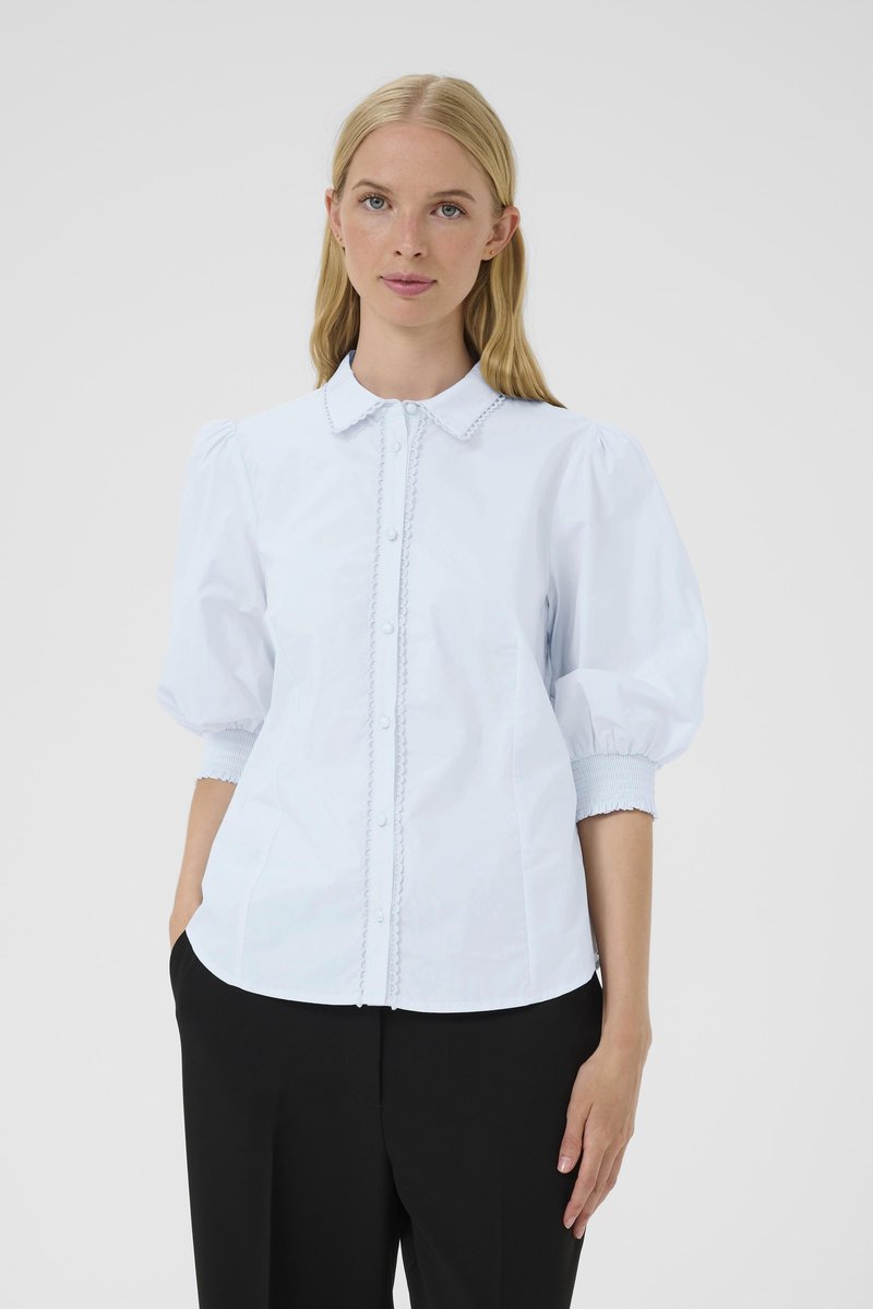 Donna bionda che indossa una camicia bianca con bottoni e maniche a sbuffo, pantaloni neri, in piedi con una mano in tasca contro uno sfondo grigio.