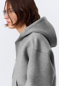Siva zip-up kapuca s široko kapuco, mehak material in subtilna tekstura. Ima zadrgo in stranske žepe.