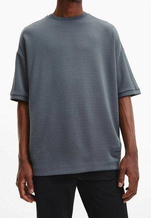 Muž stojící s uvolněnýma rukama podél těla, oblečený v volném šedém tričku s krátkými rukávy a černých kalhotách, na bílém pozadí.