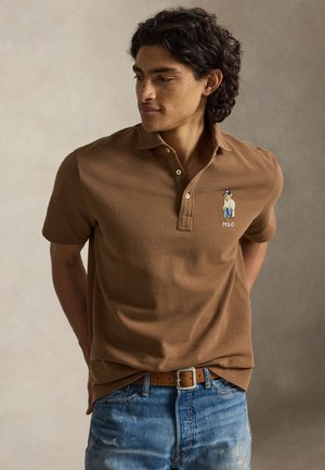 Homme aux cheveux bouclés foncés portant un polo marron avec un petit logo ours et un jean bleu, debout les mains derrière le dos.