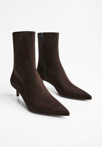O pereche de cizme de nivel ankle din suede maro închis, cu vârfuri ascuțite, tocuri joase și un fermoar lateral pe un fundal alb.