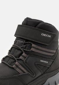 Black waterproof boot with textured upper, featuring a Velcro strap, striped accents, and logo. Designed for durability and comfort.