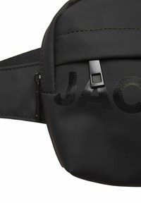 Marsupio nero realizzato in materiale morbido con il logo "JAC" lucido. Presenta una chiusura con zip e una tracolla regolabile per una vestibilità sicura.