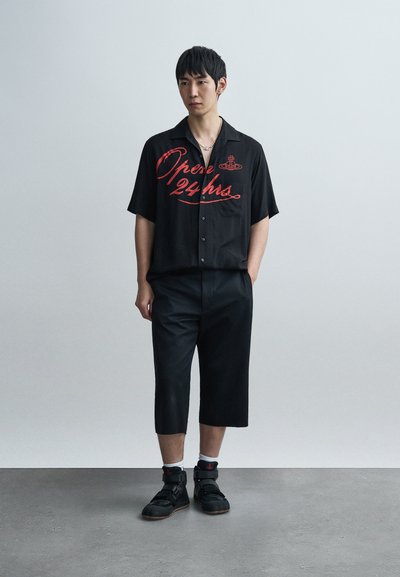 Μαύρο πουκάμισο με κοντά μανίκια και κόκκινο γράφημα "Ανοικτά 24 ώρες", συνδυασμένο με μαύρο cropped παντελόνι και μαύρα αθλητικά παπούτσια με ρυθμιζόμενα λουράκια.