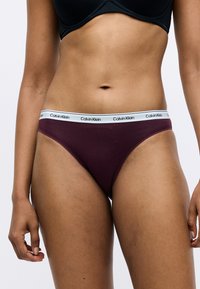 Calvin Klein bordó pamutszövött tanga, fehér logós derékpánttal; sima felület és alacsony fazon a kényelemért és minimális takarásért.