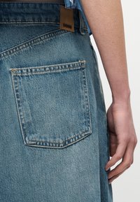 Κοντινή λήψη της πίσω τσέπης μπλε τζιν με καφέ δερμάτινη ετικέτα BOSS και το χέρι ενός ατόμου που ακουμπά δίπλα στο ισχίο.