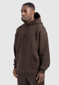 Sudadera con capucha marrón hecha de tela suave. Presenta un bolsillo frontal y detalles de logotipo en relieve. Corte holgado con puños acanalados.