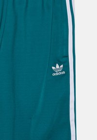 Pantalones Adidas en verde azulado con franjas blancas en los lados y el logotipo bordado de trefoil de Adidas cerca del bolsillo en la pierna derecha.