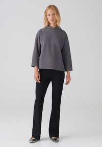 Felpa grigia con cappuccio, dal design squadrato e maniche ampie, abbinata a pantaloni neri a zampa e scarpe flat metalliche. Sfondo semplice.