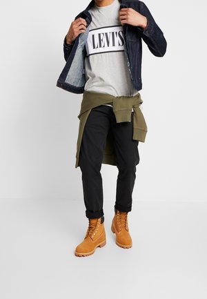 Grauer Levi's Sweatshirt mit schwarzem Logo, unter einer dunklen Jeansjacke getragen, kombiniert mit schwarzen Hosen und tanfarbenen Schnürstiefeln. Olivgrünes Sweatshirt um die Taille gebunden.