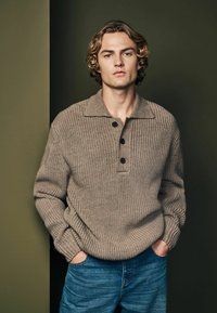 Pull en beige tricoté avec un col et trois boutons noirs, associé à un jean en denim bleu. Le modèle se tient devant un fond vert foncé.